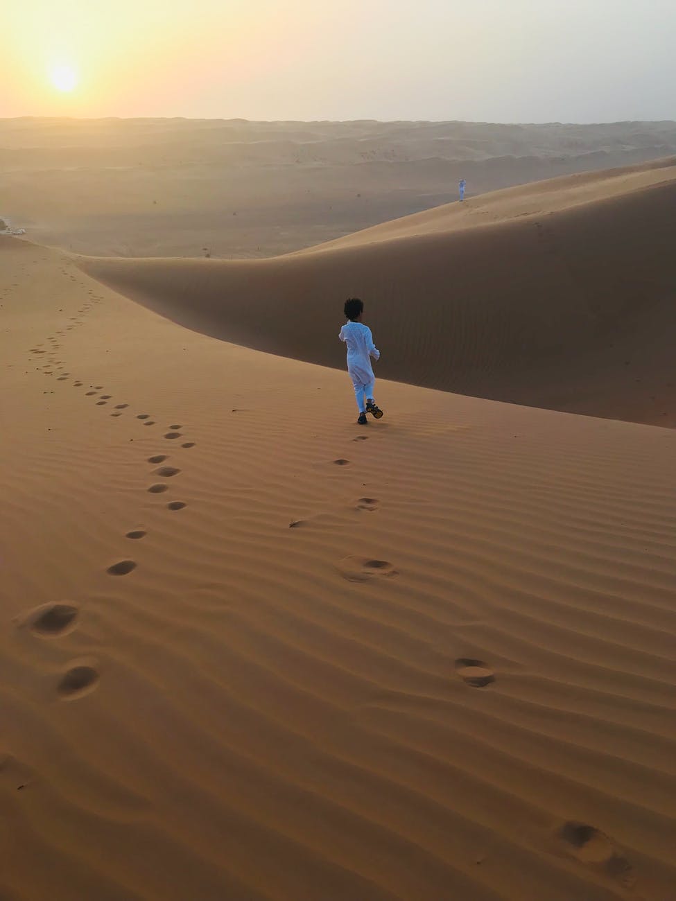 back view photo of muslim boy in white thobe walking alone on desert sand during golden hour